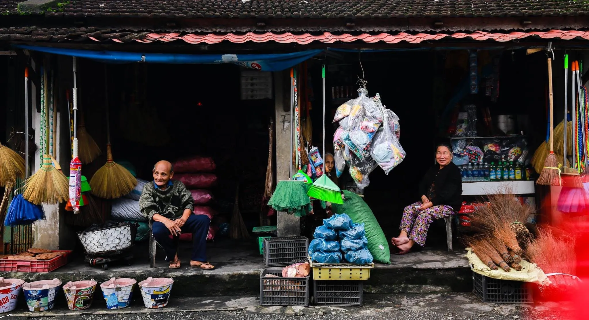 Two elderly shopkeepers rest in front of a shop hung with brooms in Bao Vinh ancient town